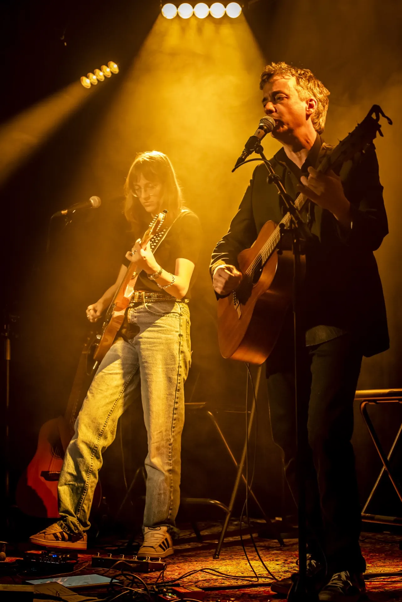 Duo debout, guitare électrique et acoustique, faisceau doré tombant depuis les projecteurs.
