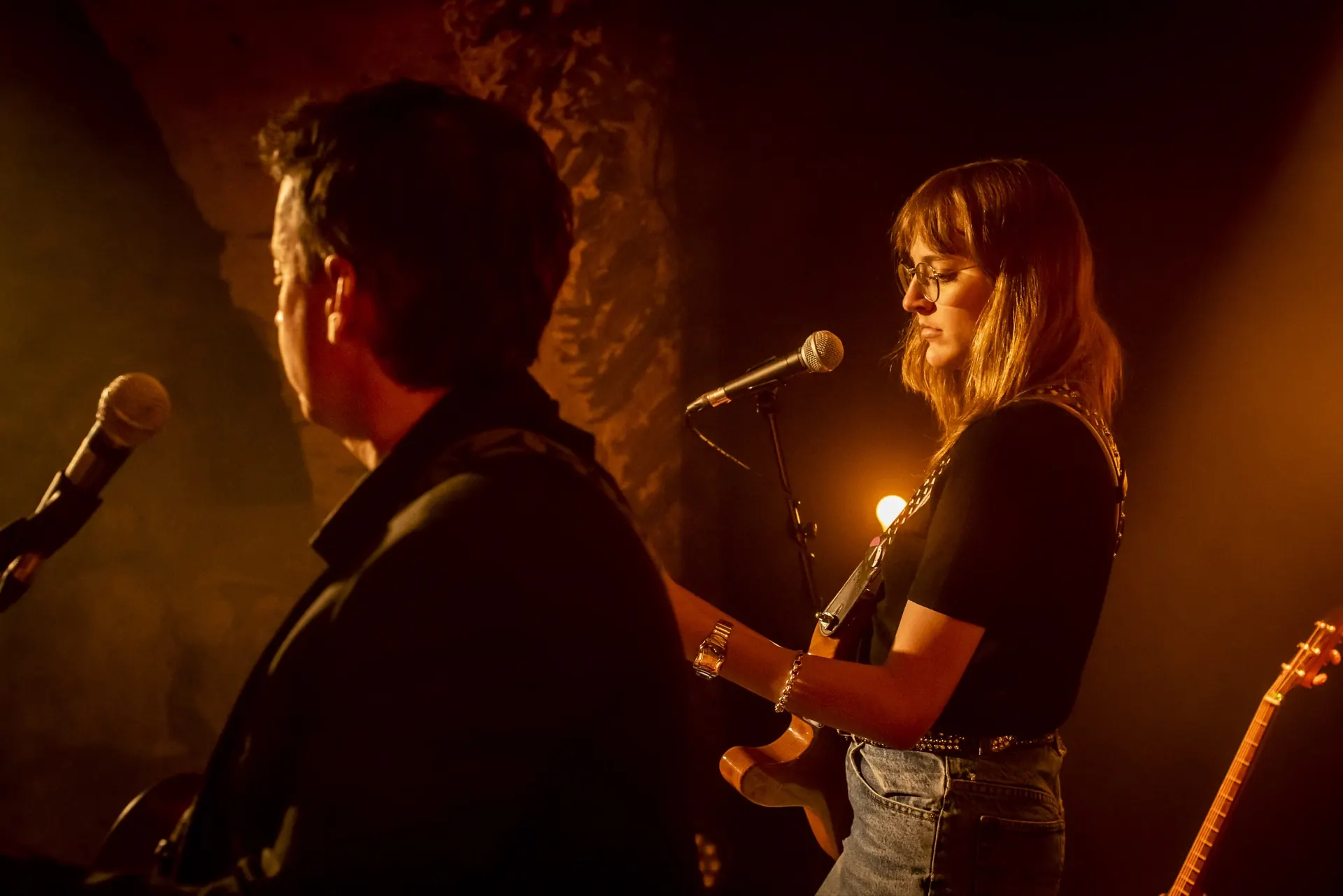 Chanteuse au micro à droite, guitariste de dos au premier plan, lumière chaude.