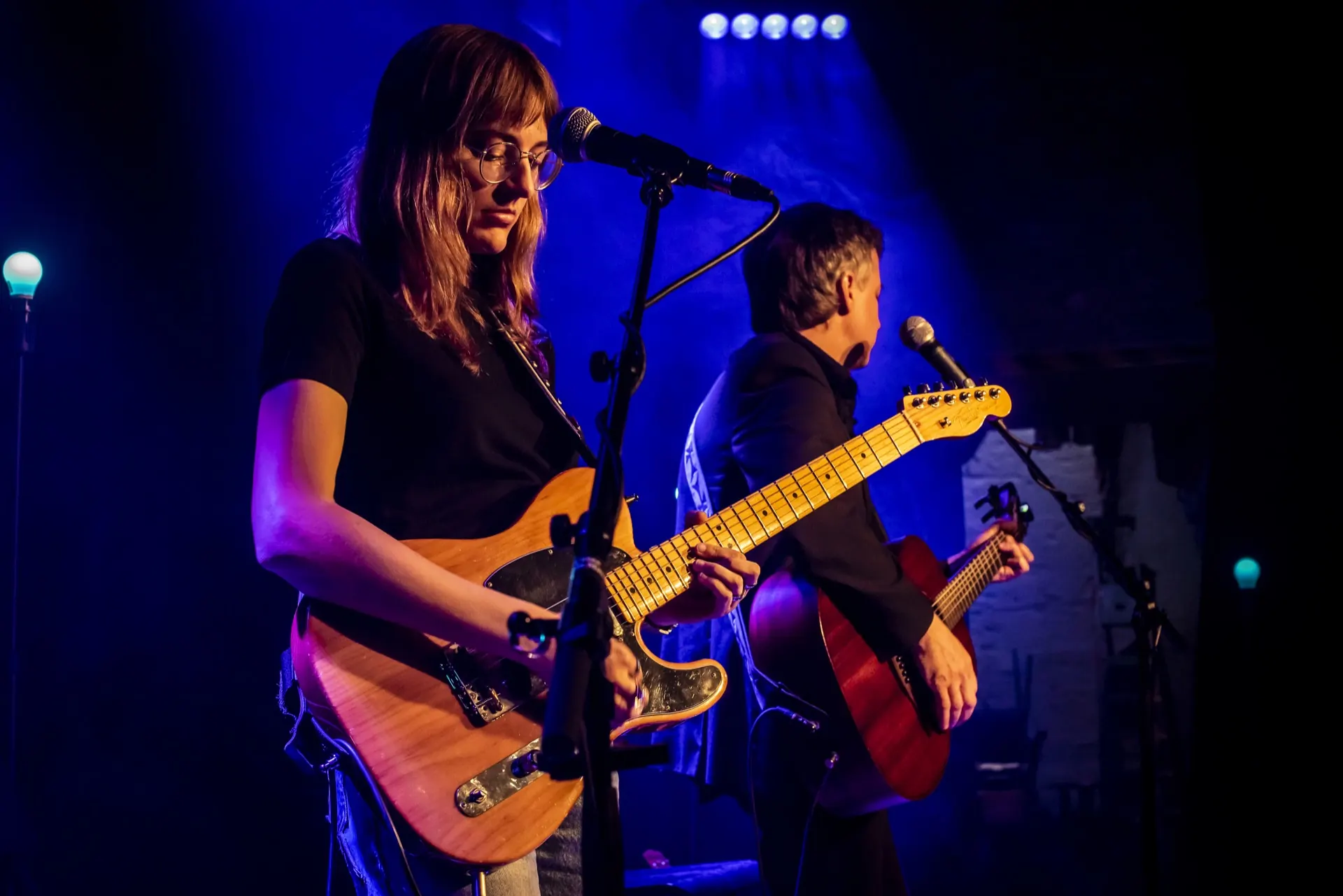 Guitariste à la Telecaster au micro, un second musicien flou derrière, lumière bleue.