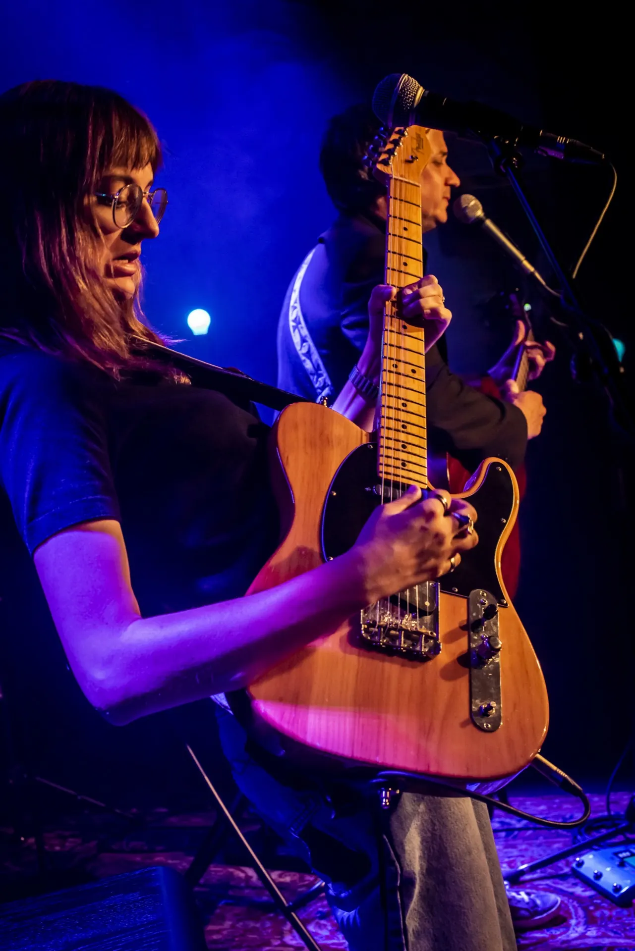 Profil d'une guitariste à la Telecaster, musicien au micro derrière, ambiance bleutée.
