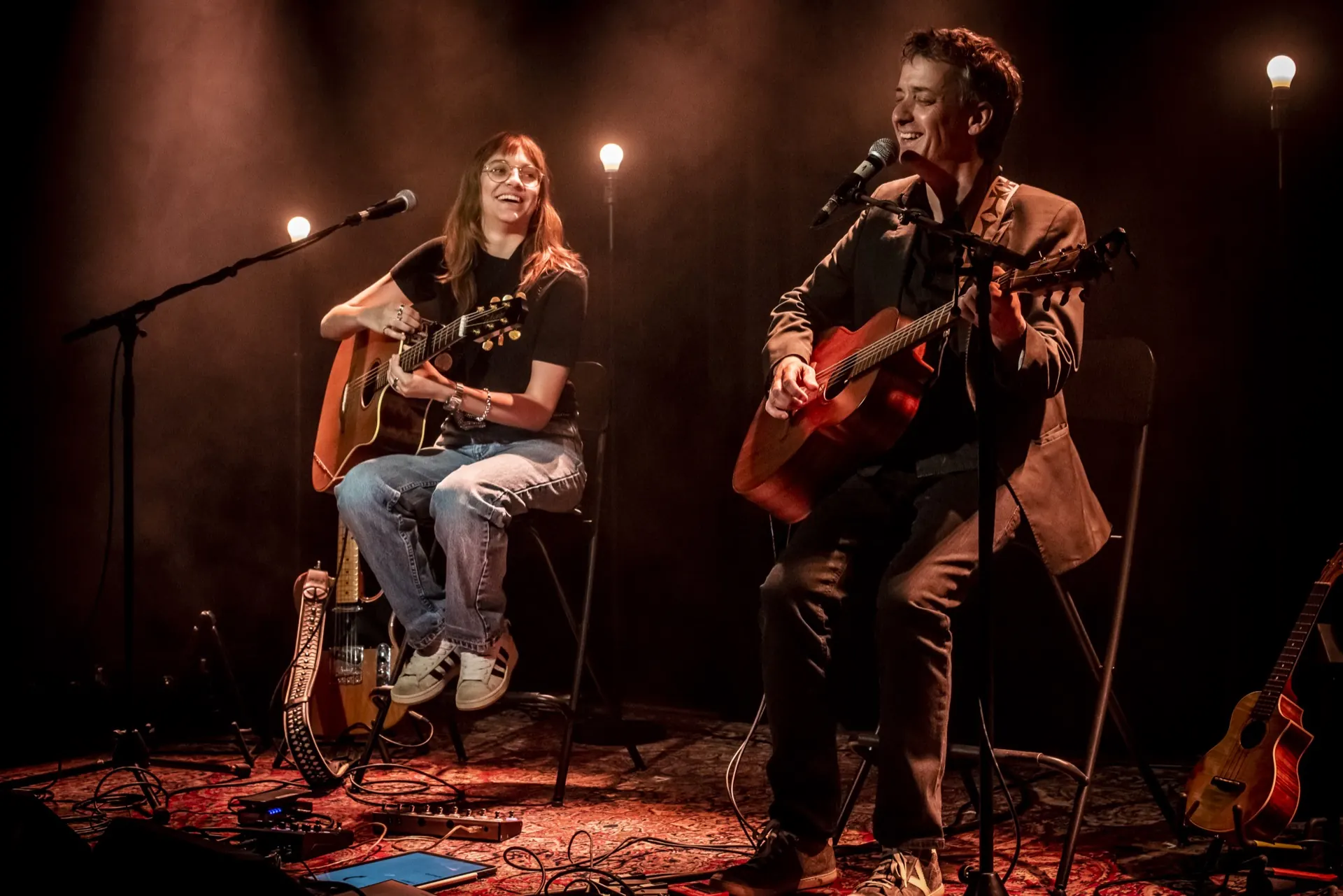 Deux musiciens assis souriant, guitares acoustiques, lumière ambrée, tapis et câbles au sol.