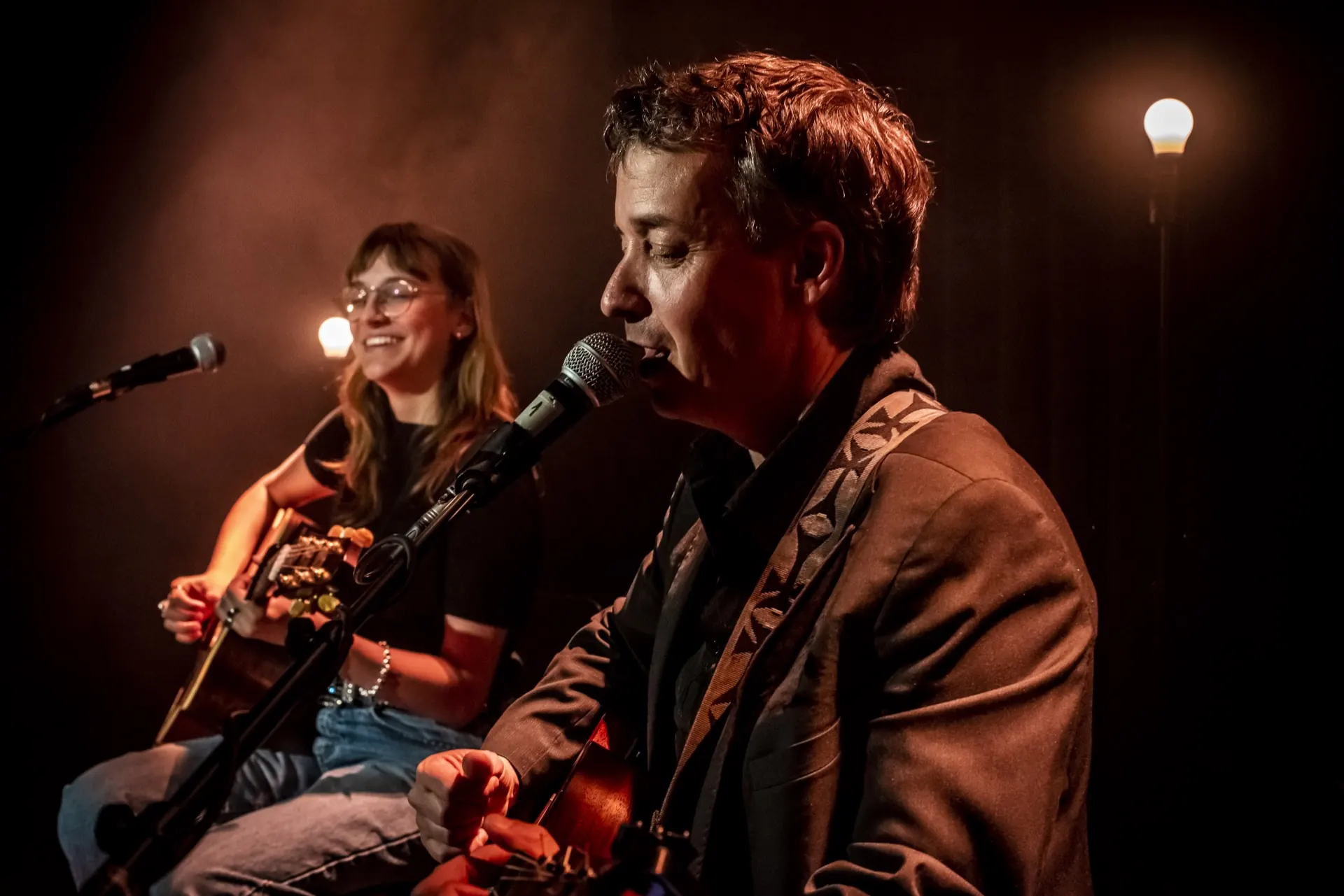 Chanteur de profil au micro, musicienne souriante en retrait, lumière brune feutrée.