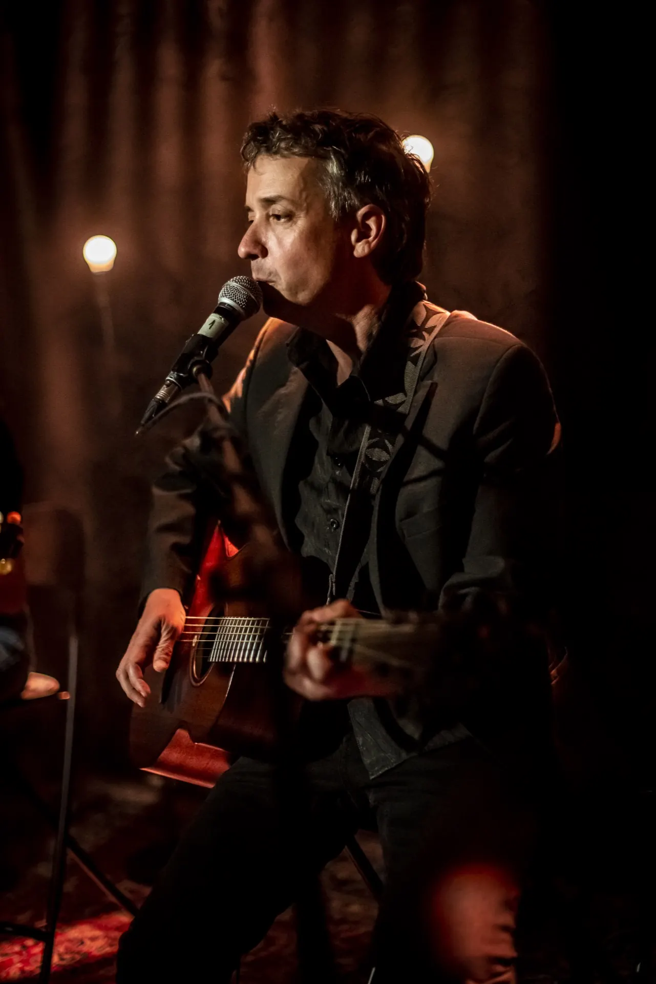 Chanteur seul assis au micro, guitare acoustique, ampoules allumées dans le fond sombre.