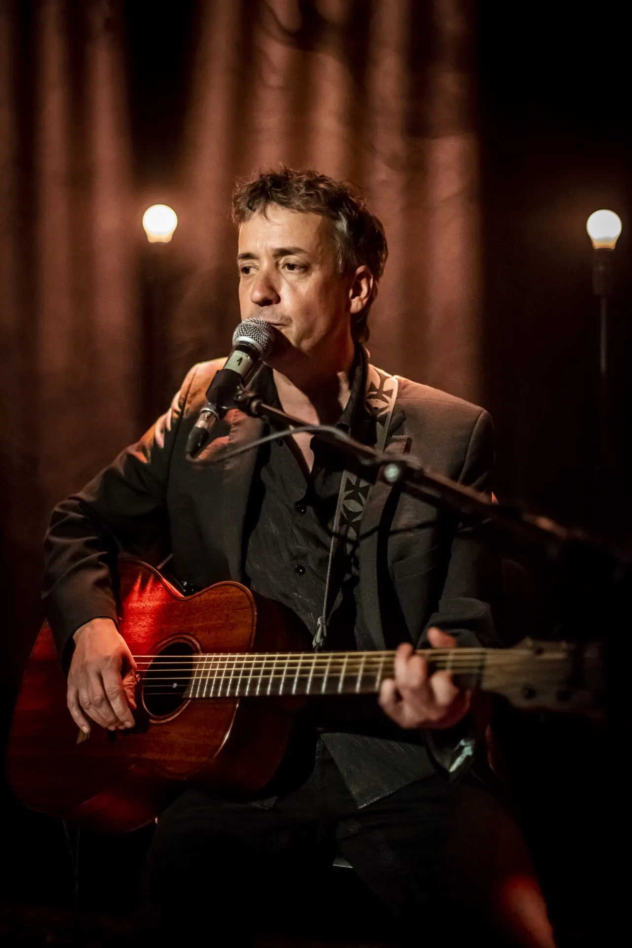 Musicien assis chantant, guitare rouge sur les genoux, deux lampes en arrière-plan.