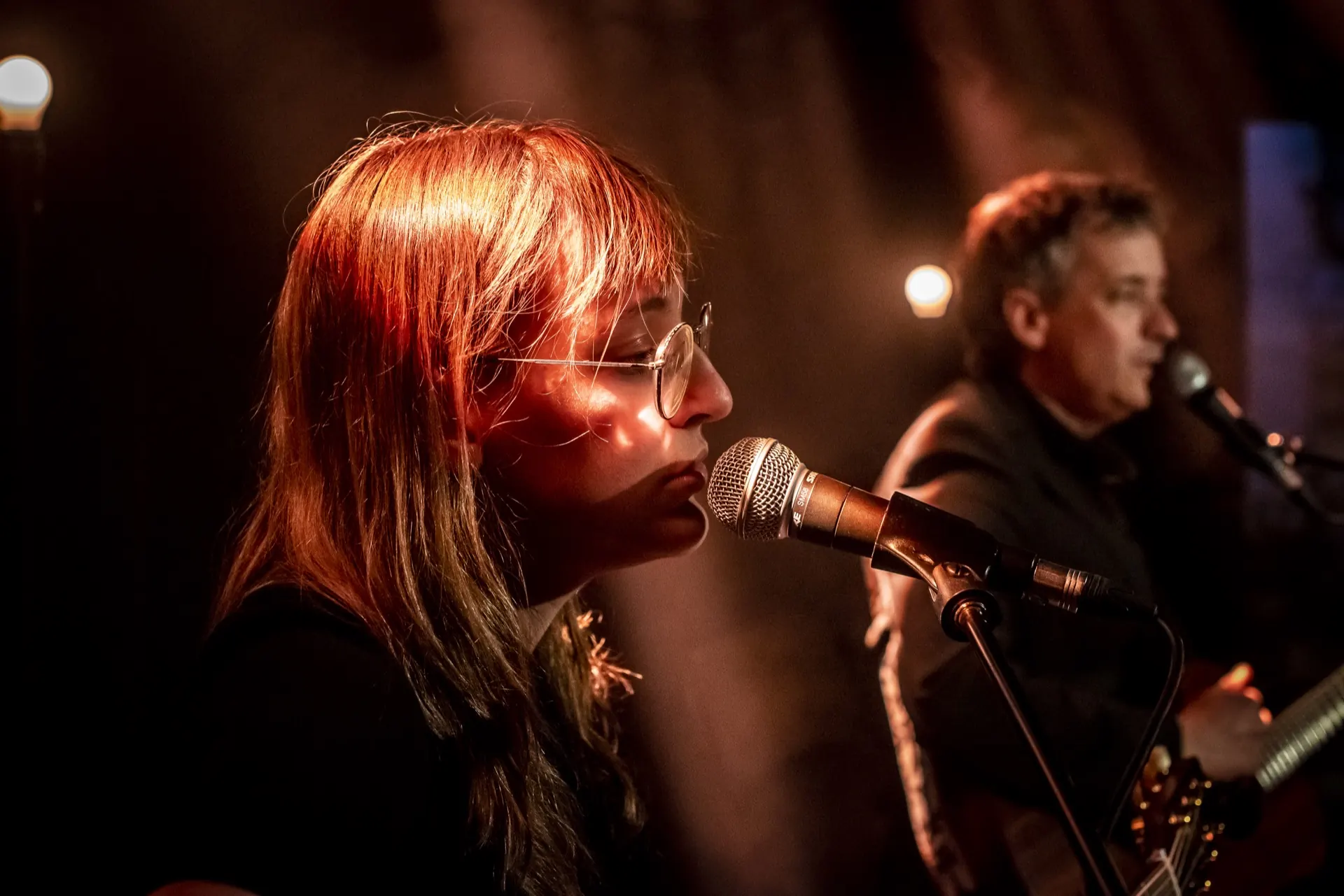 Chanteuse de profil au micro, chevelure éclairée de rouge, guitariste dans l'ombre.