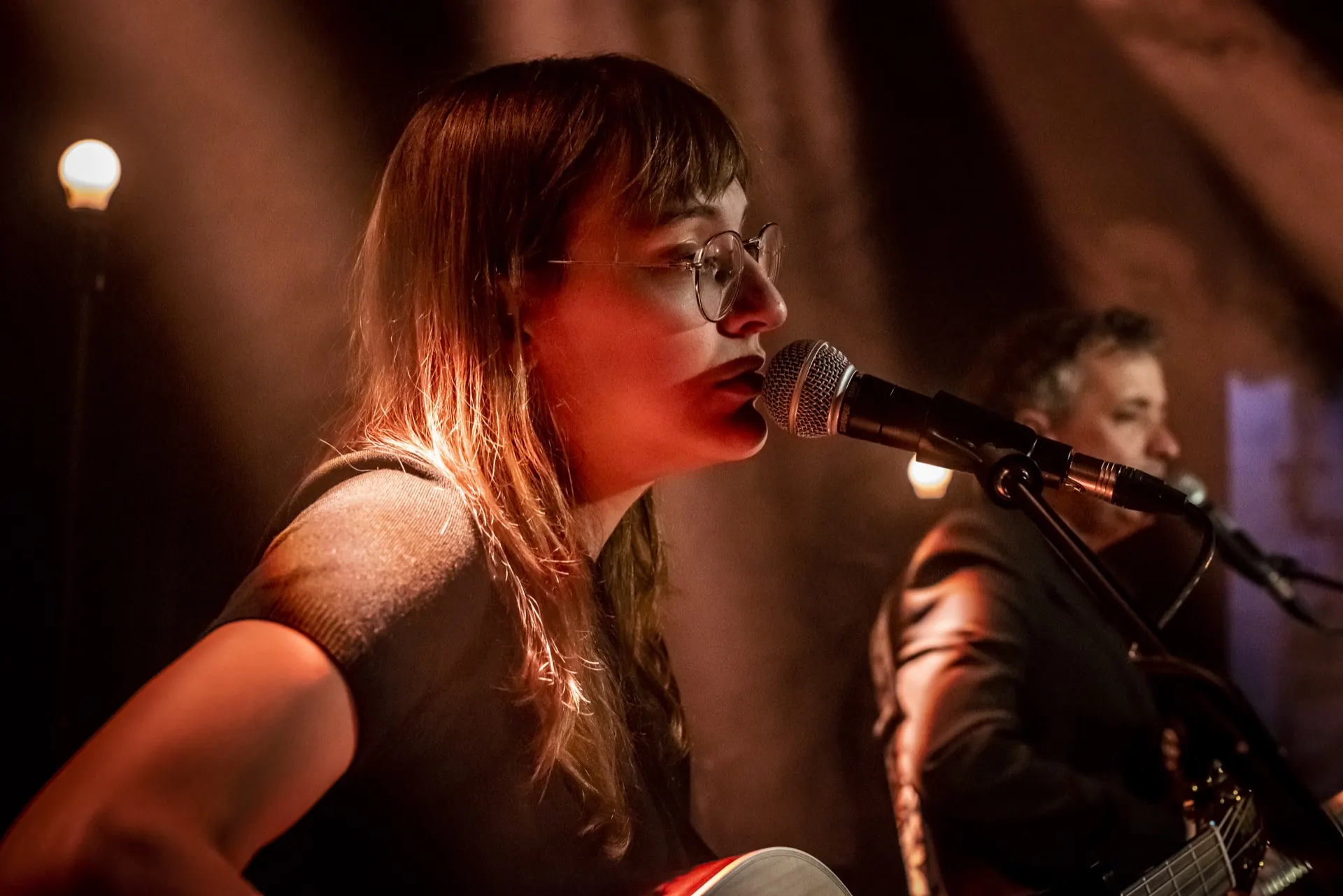 Portrait rapproché d'une chanteuse à lunettes au micro, musicien flou, lumière chaude.