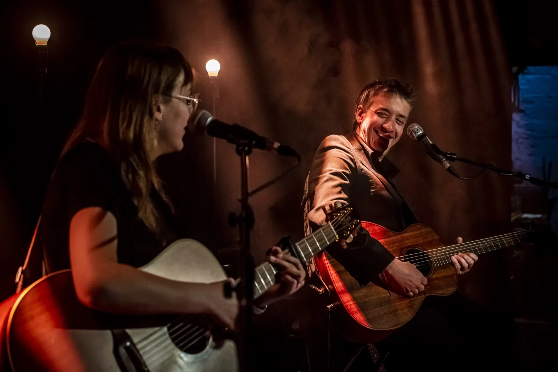 Chanteuse à gauche au micro, guitariste riant à droite, guitares acoustiques, lumière chaude.