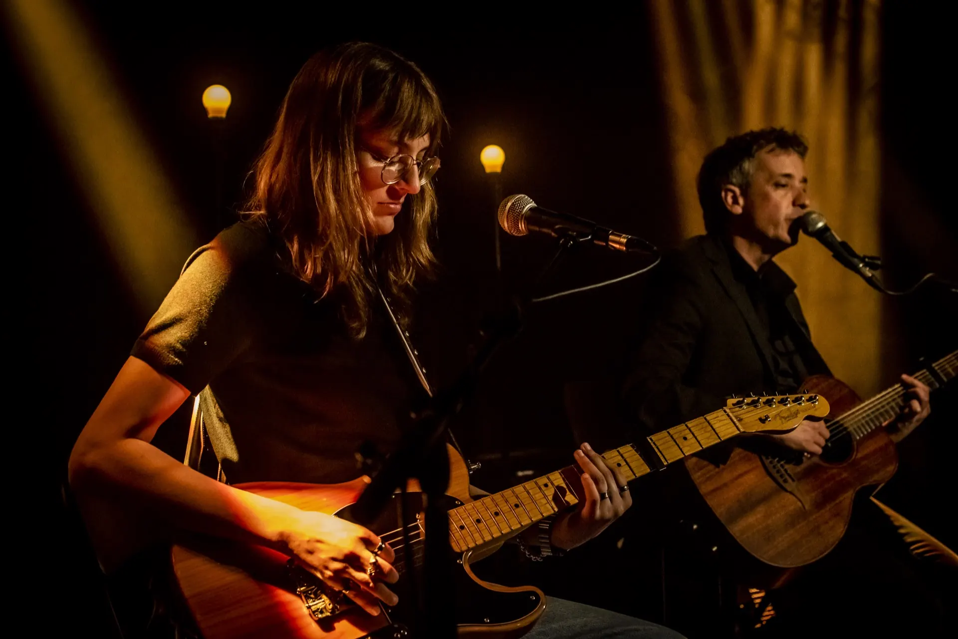 Guitare électrique au premier plan, chanteur avec acoustique à droite, ampoules et fumée ambrée.