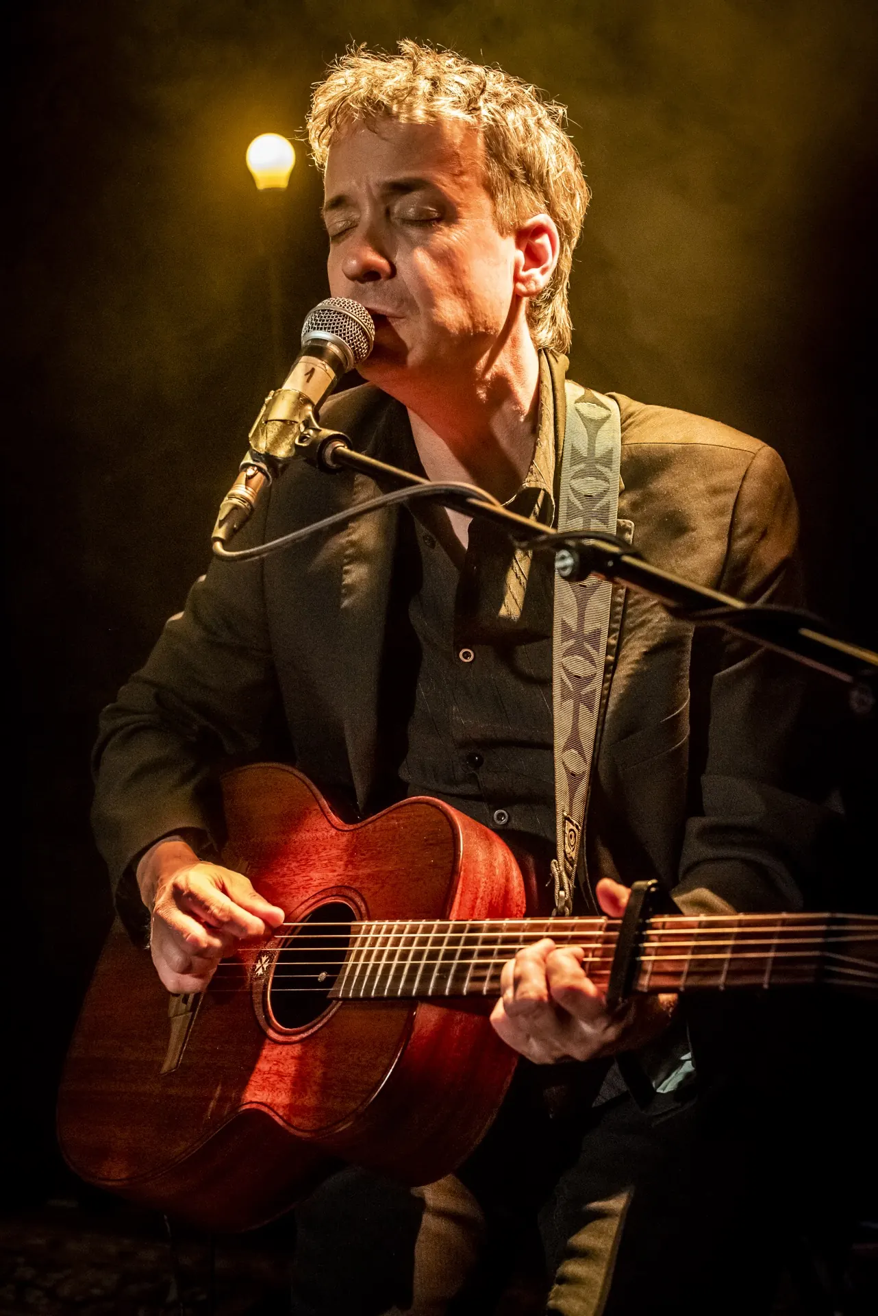 Chanteur au micro avec guitare acoustique brune, halo ambré et ampoule en arrière-plan.