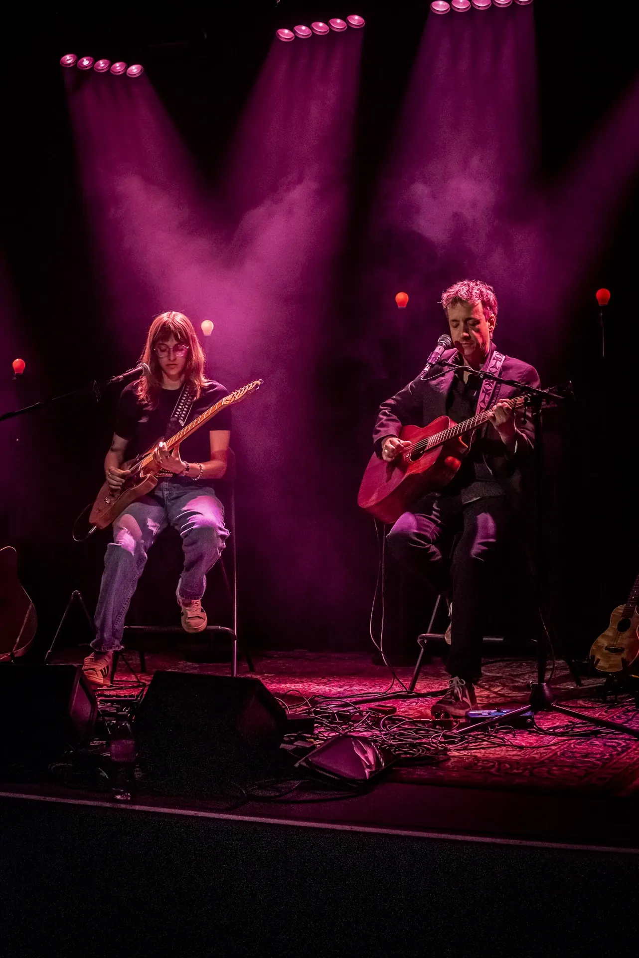 Duo assis en plan large, guitare électrique et acoustique, projecteurs et fumée magenta.
