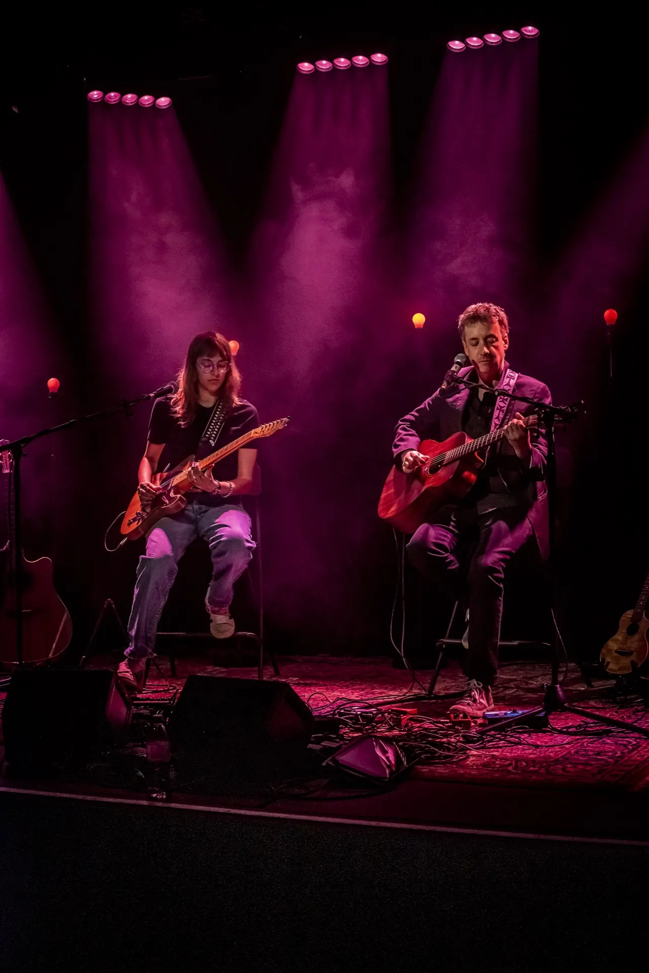 Deux musiciens assis, lumière rose et magenta, faisceaux de projecteurs, rideau au fond.