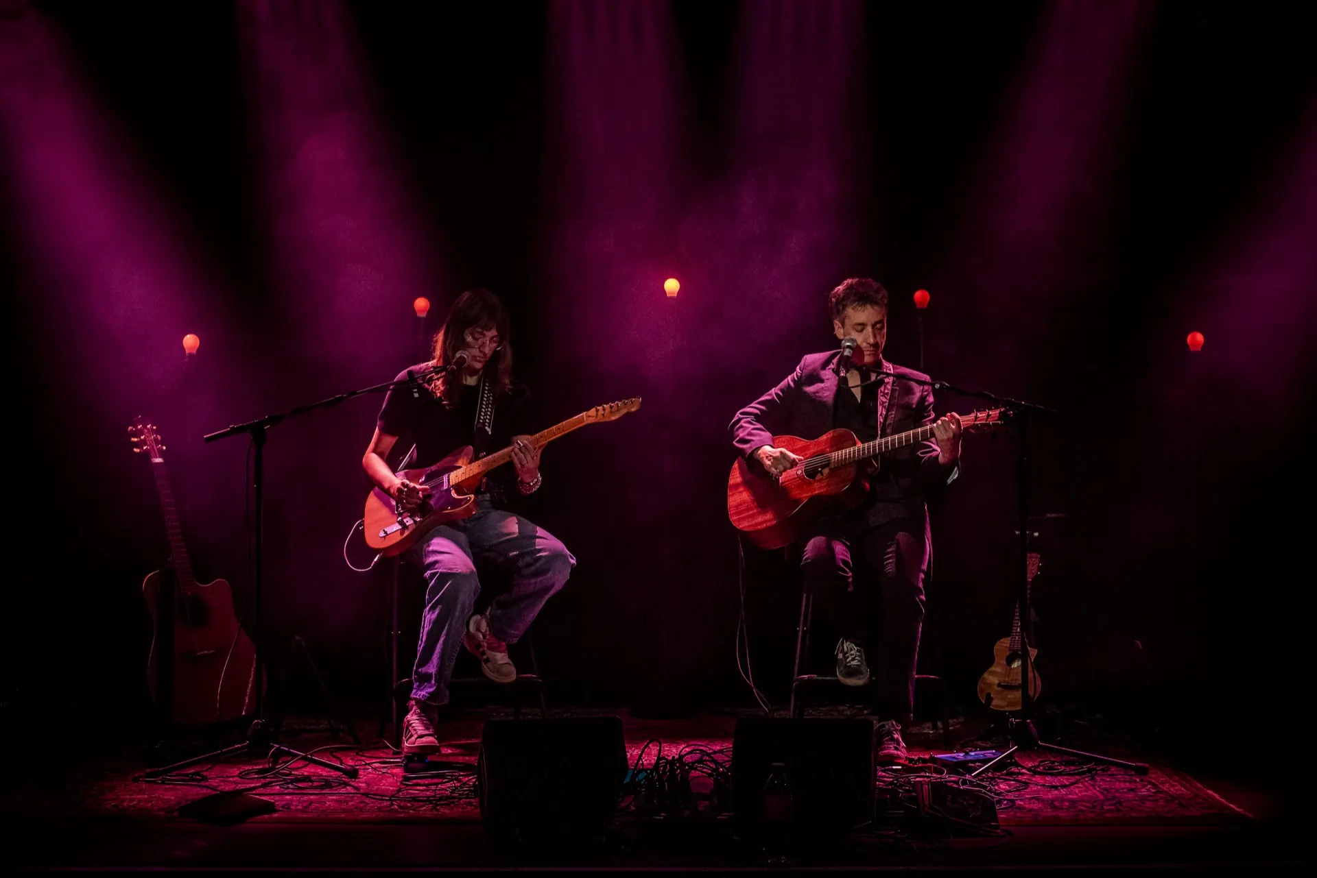 Deux guitaristes assis, l'un électrique l'autre acoustique, plan large baigné de magenta profond.