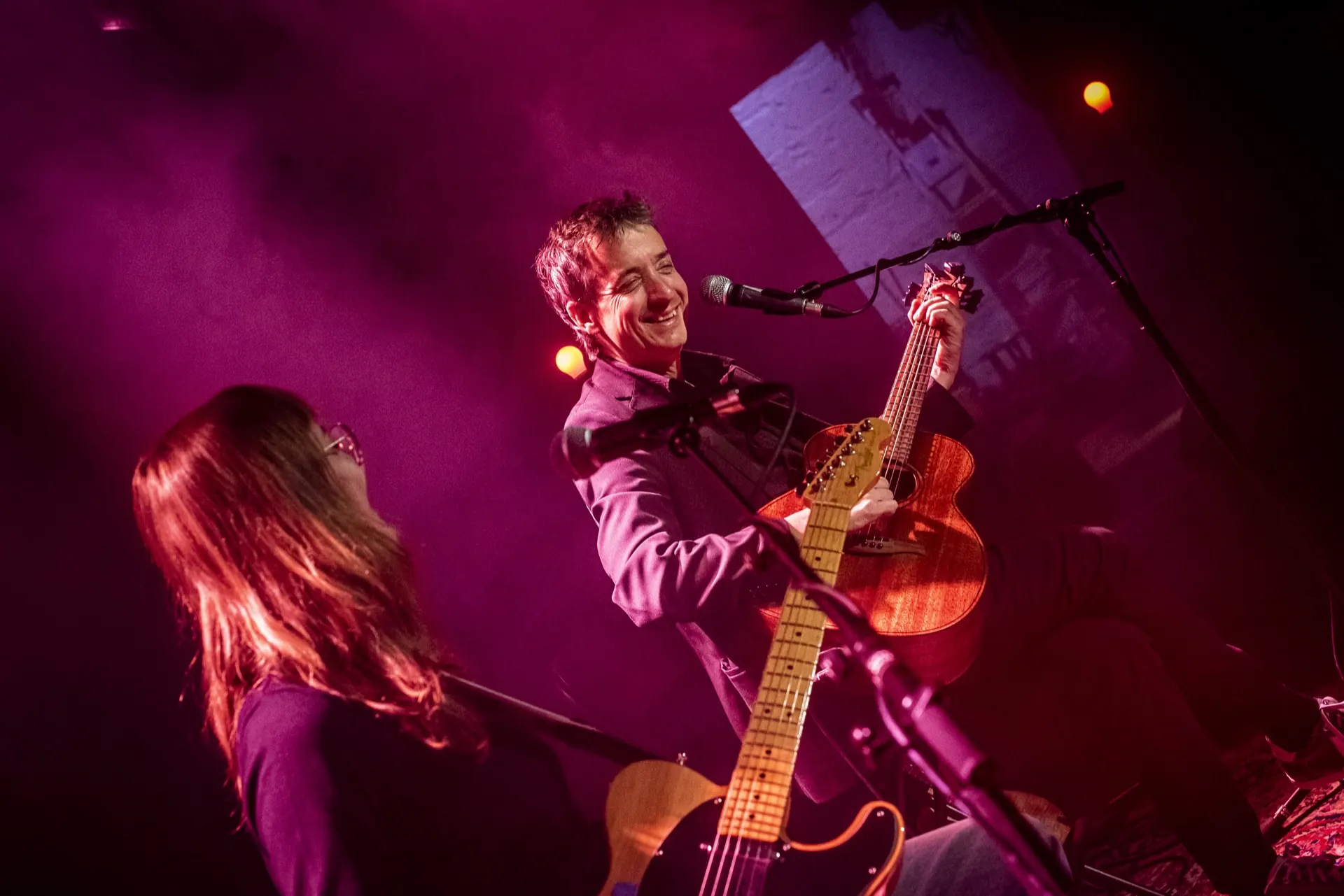 Chanteur souriant au micro, musicienne floue au premier plan, pupitre et lumière magenta.