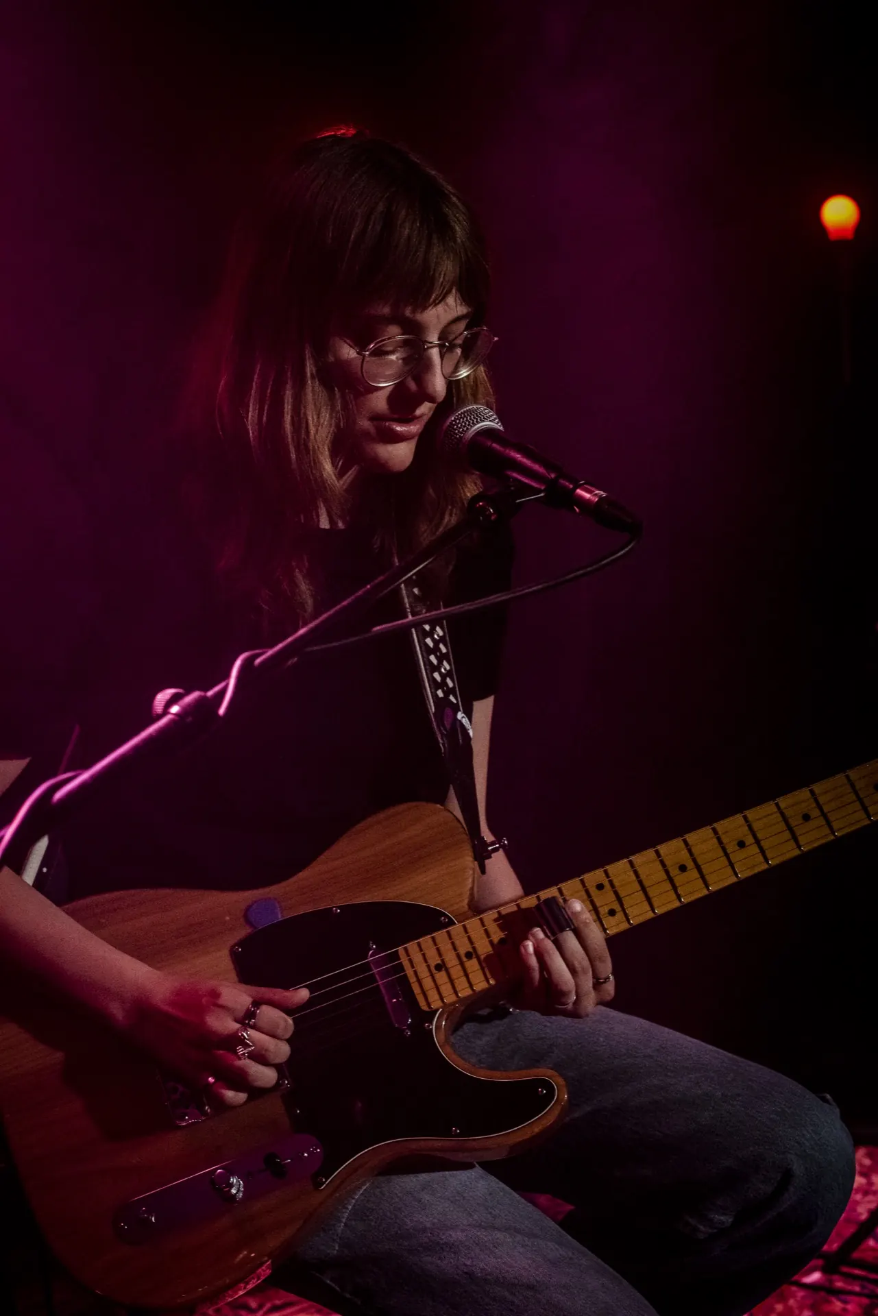 Guitariste assise à la Telecaster au micro, lunettes, dégradé violet et magenta.
