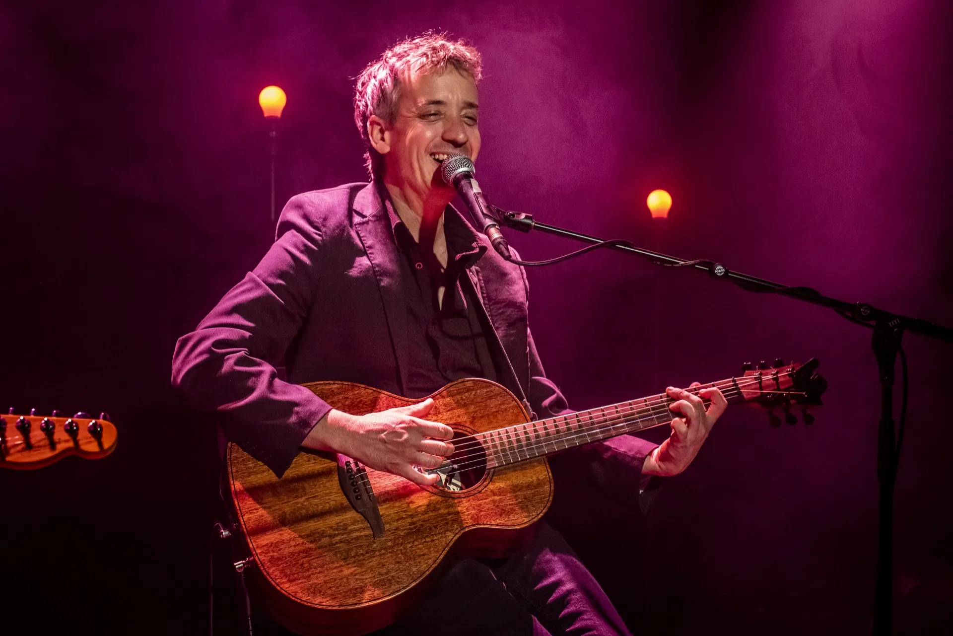 Chanteur assis, large sourire, guitare acoustique, ampoules floues dans un fond magenta.