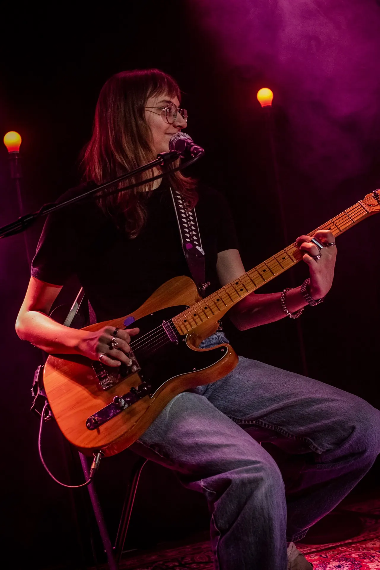 Guitariste assise à la Telecaster naturelle chantant au micro, lunettes, lumière rose.