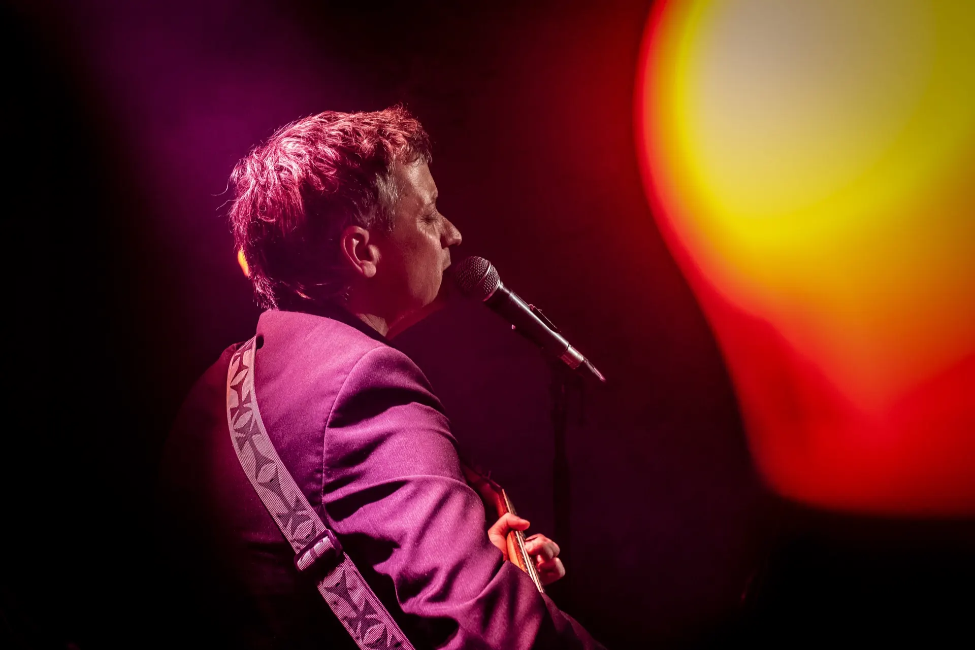 Chanteur de profil au micro, veste violette, gros halo orangé sur la droite.