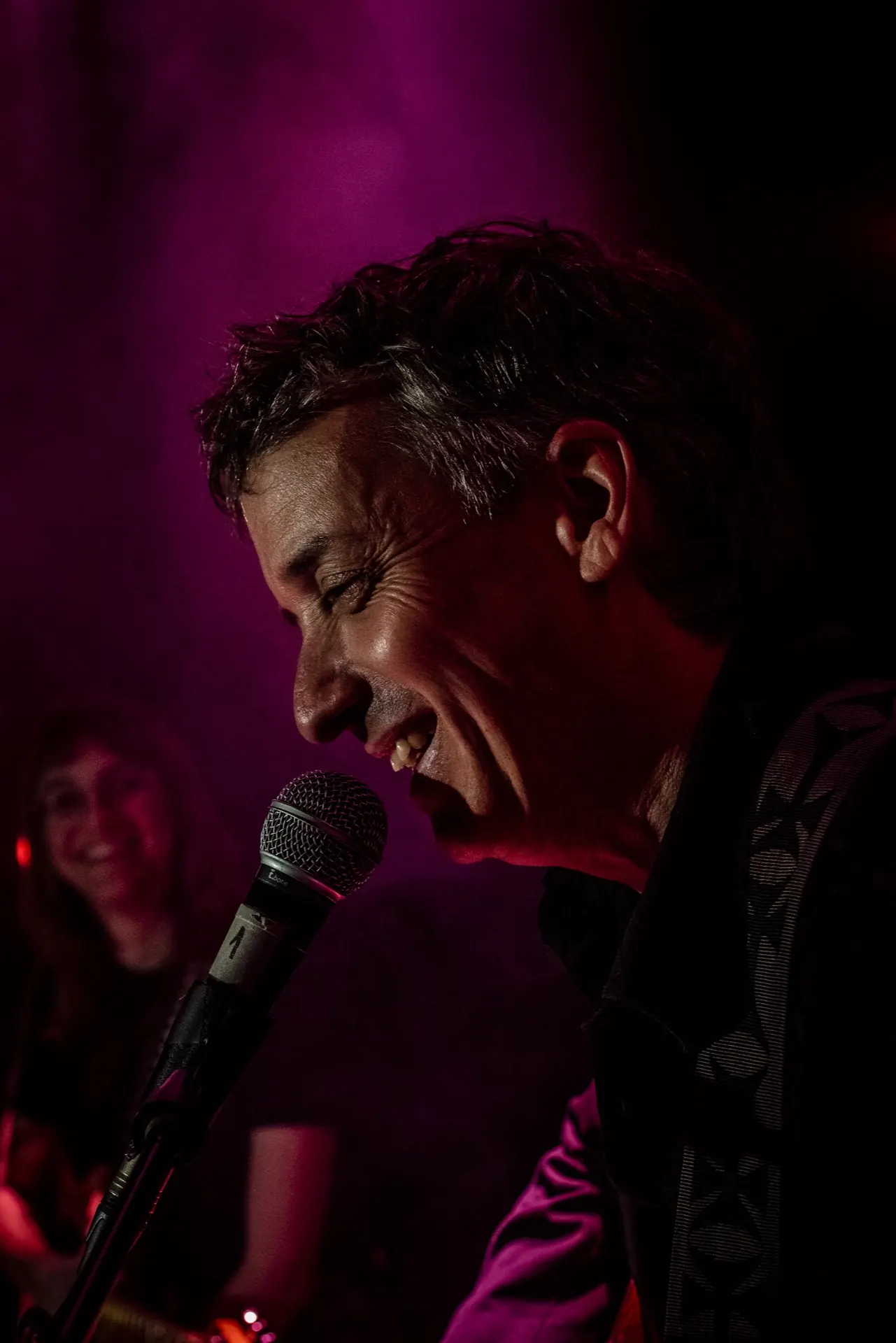 Gros plan d'un chanteur riant au micro, musicienne souriante et floue à gauche.