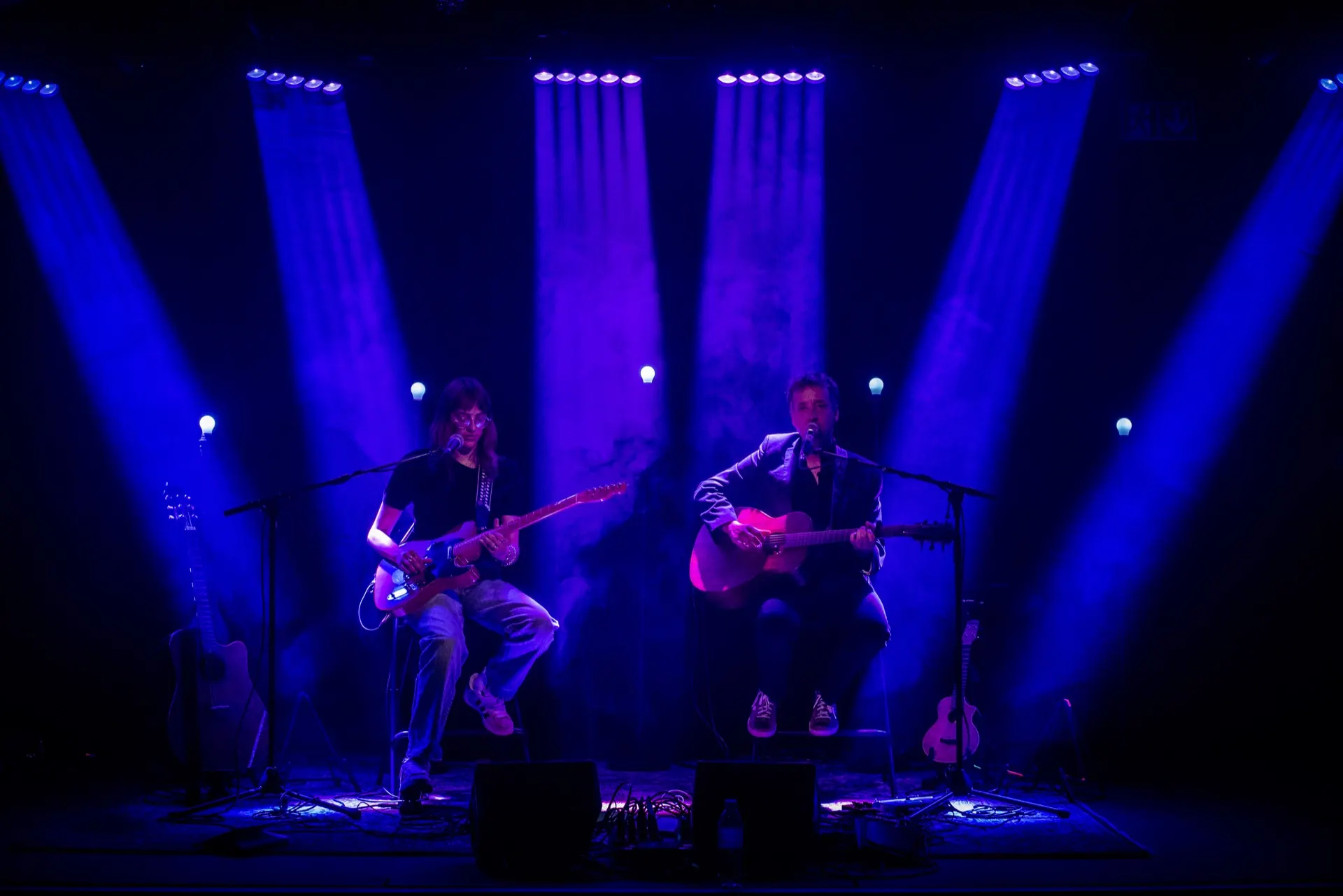 Duo assis en plan large, faisceaux bleus et rideau clair, guitares électrique et acoustique.