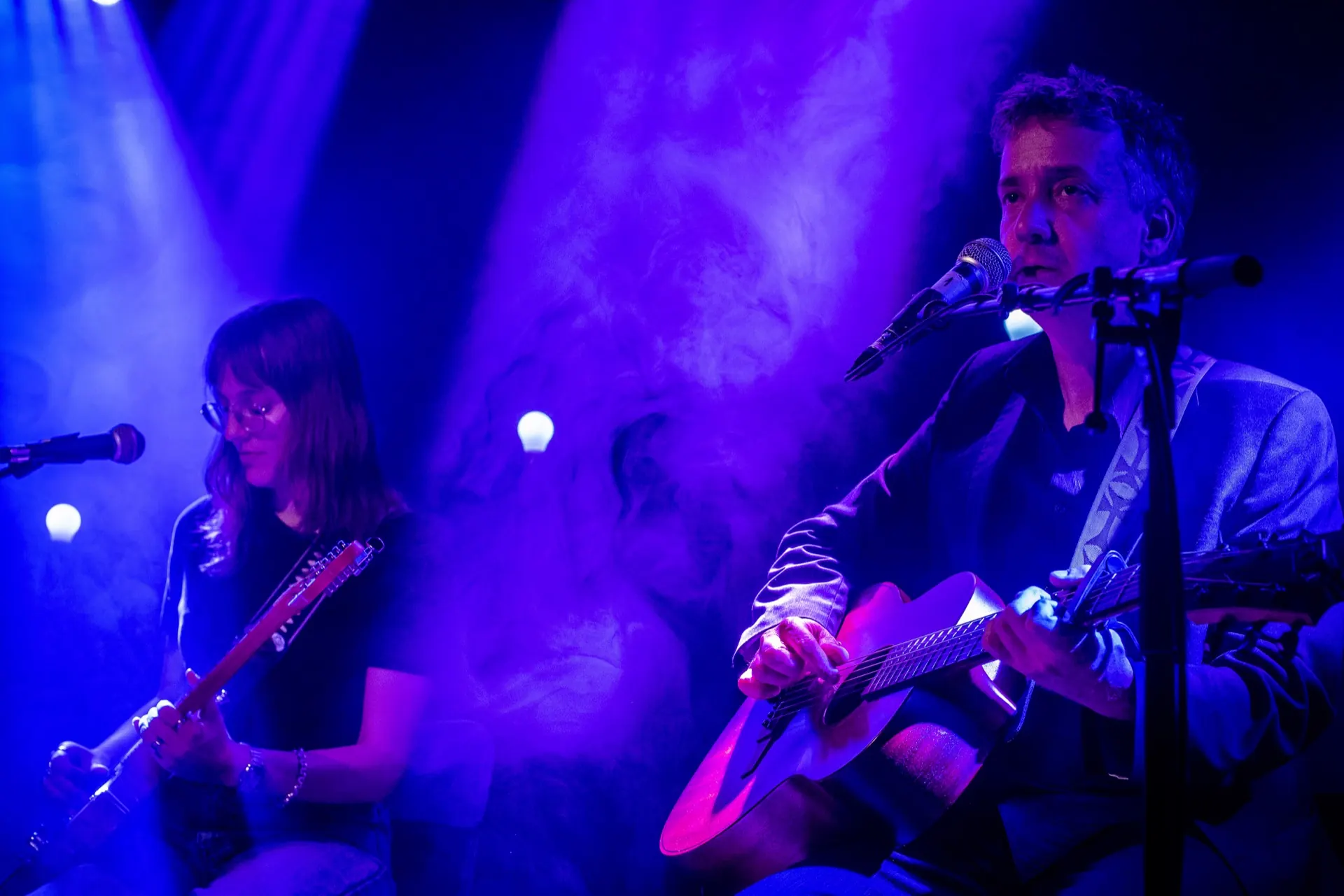Guitariste électrique à gauche, chanteur à l'acoustique à droite, fumée bleue diffuse.