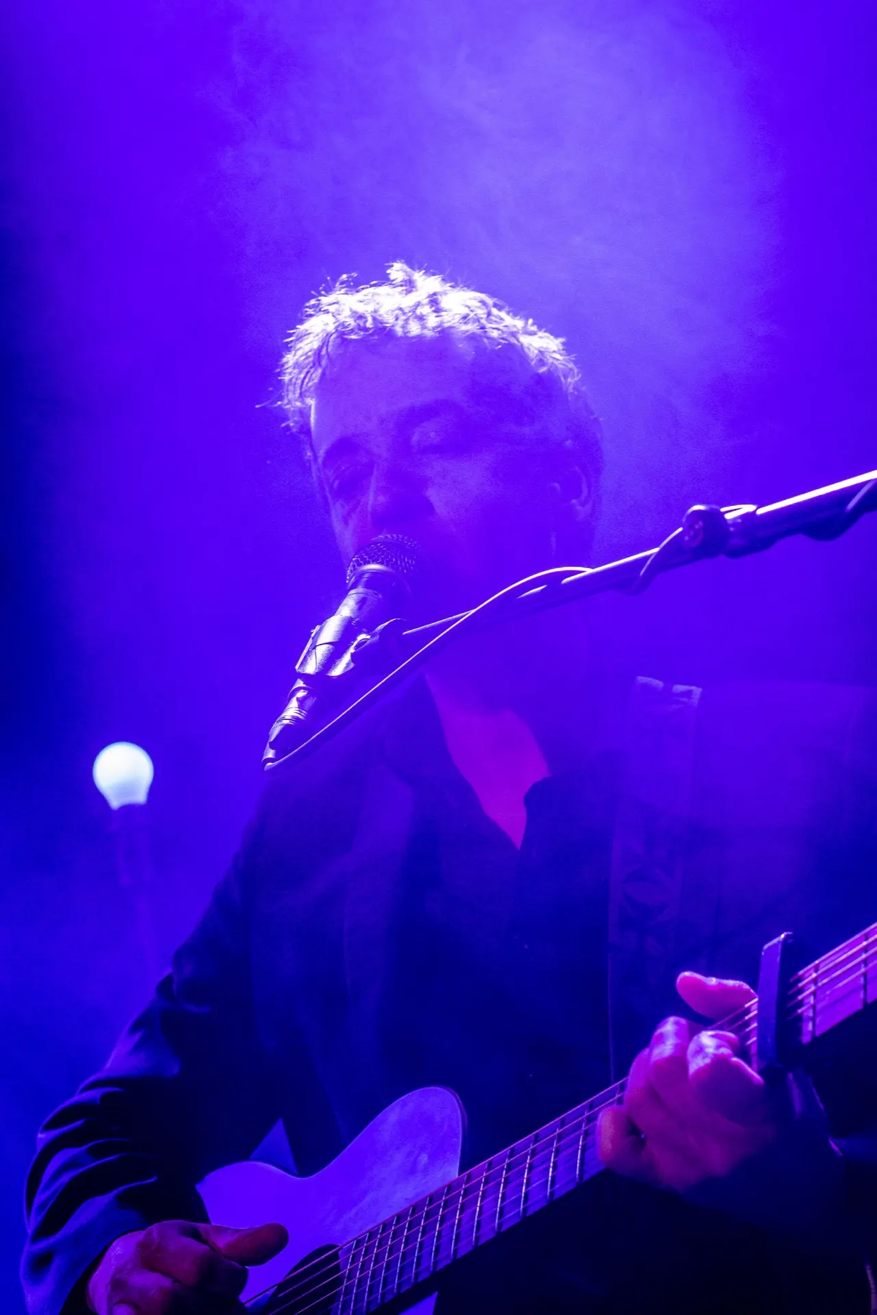 Chanteur au micro avec guitare électrique claire, halo bleu-violet et fumée.