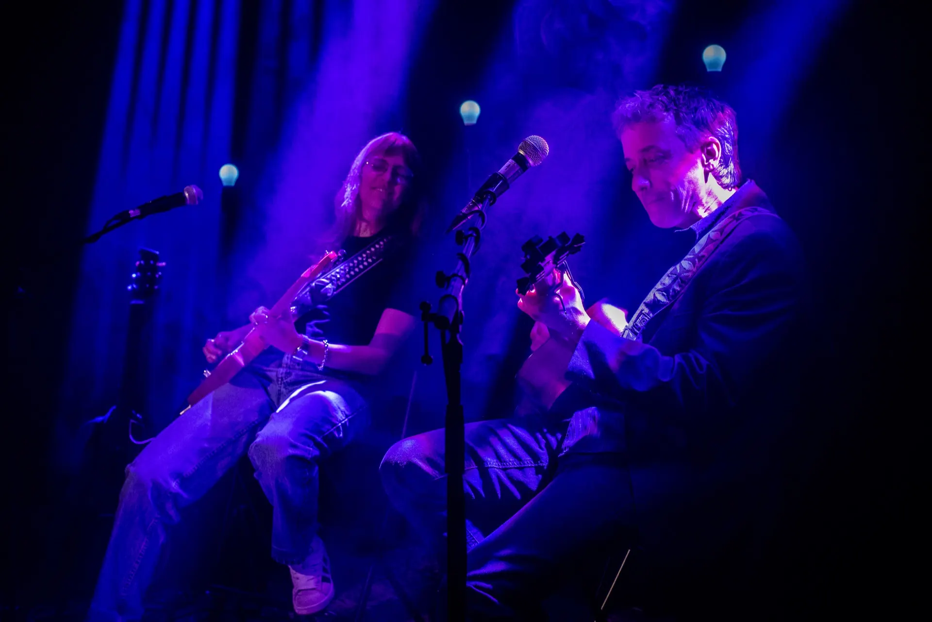 Deux guitaristes assis, l'un électrique l'autre acoustique, lumière bleu-violet et fumée.