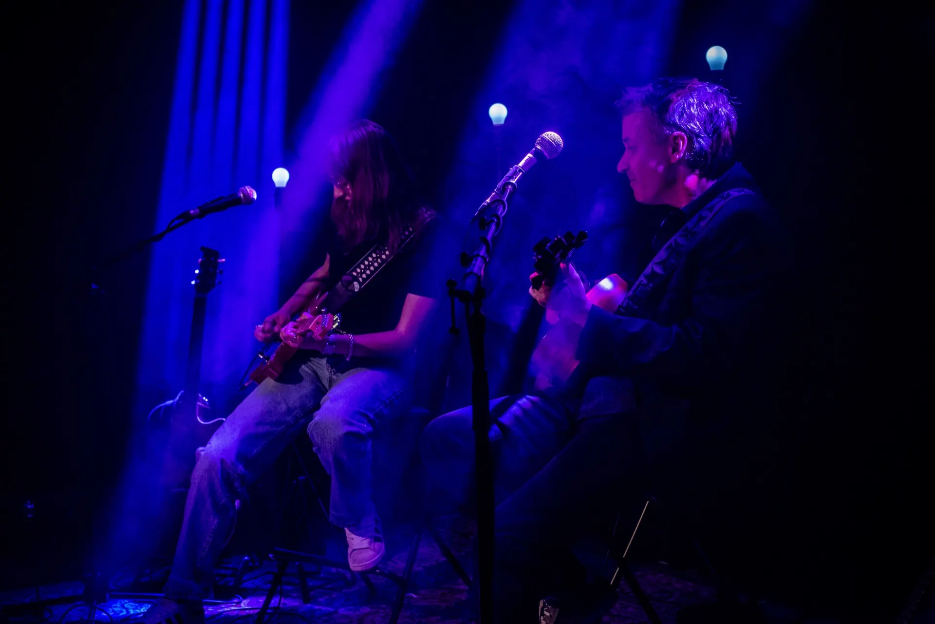 Duo assis aux micros, guitare électrique et acoustique, ampoules bleutées en arrière-plan.
