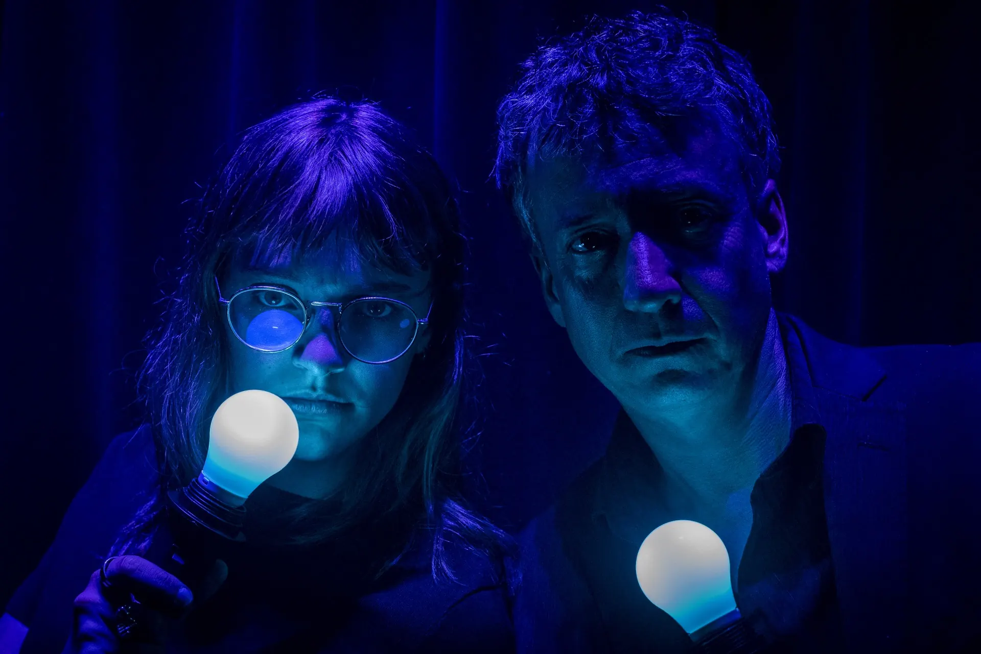 Portrait rapproché du duo tenant chacun une ampoule allumée, lumière bleue, rideau sombre.