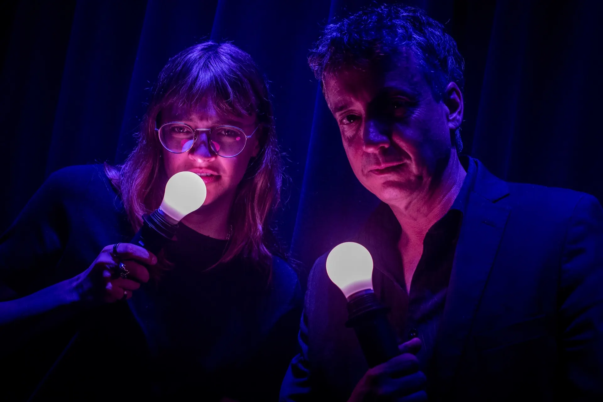 Deux visages proches éclairés par des ampoules tenues sous le menton, teintes bleu-violet.