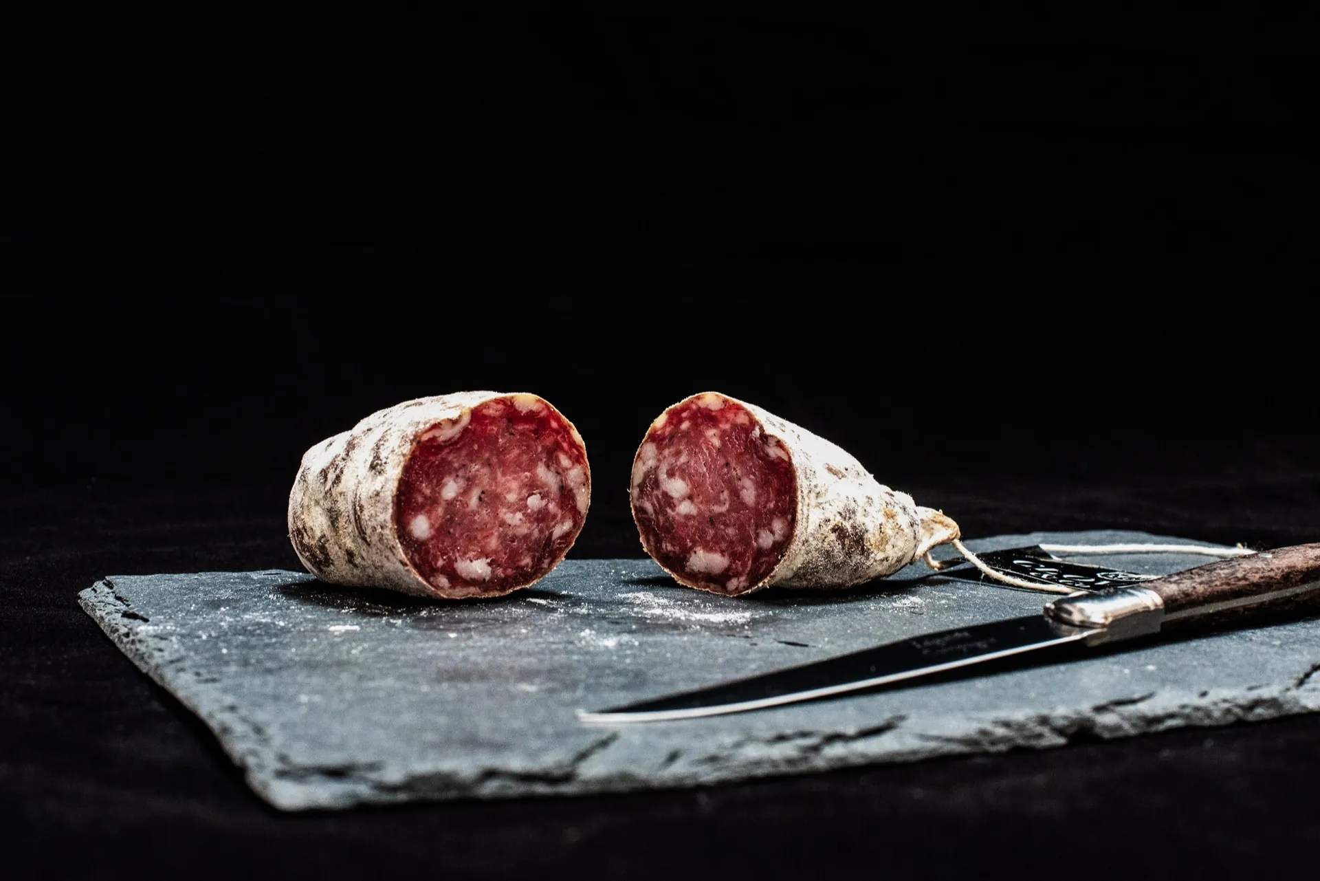 Saucisson sec tranché sur une ardoise, couteau à côté — image d’ambiance pour la Boucherie du St-Bernard